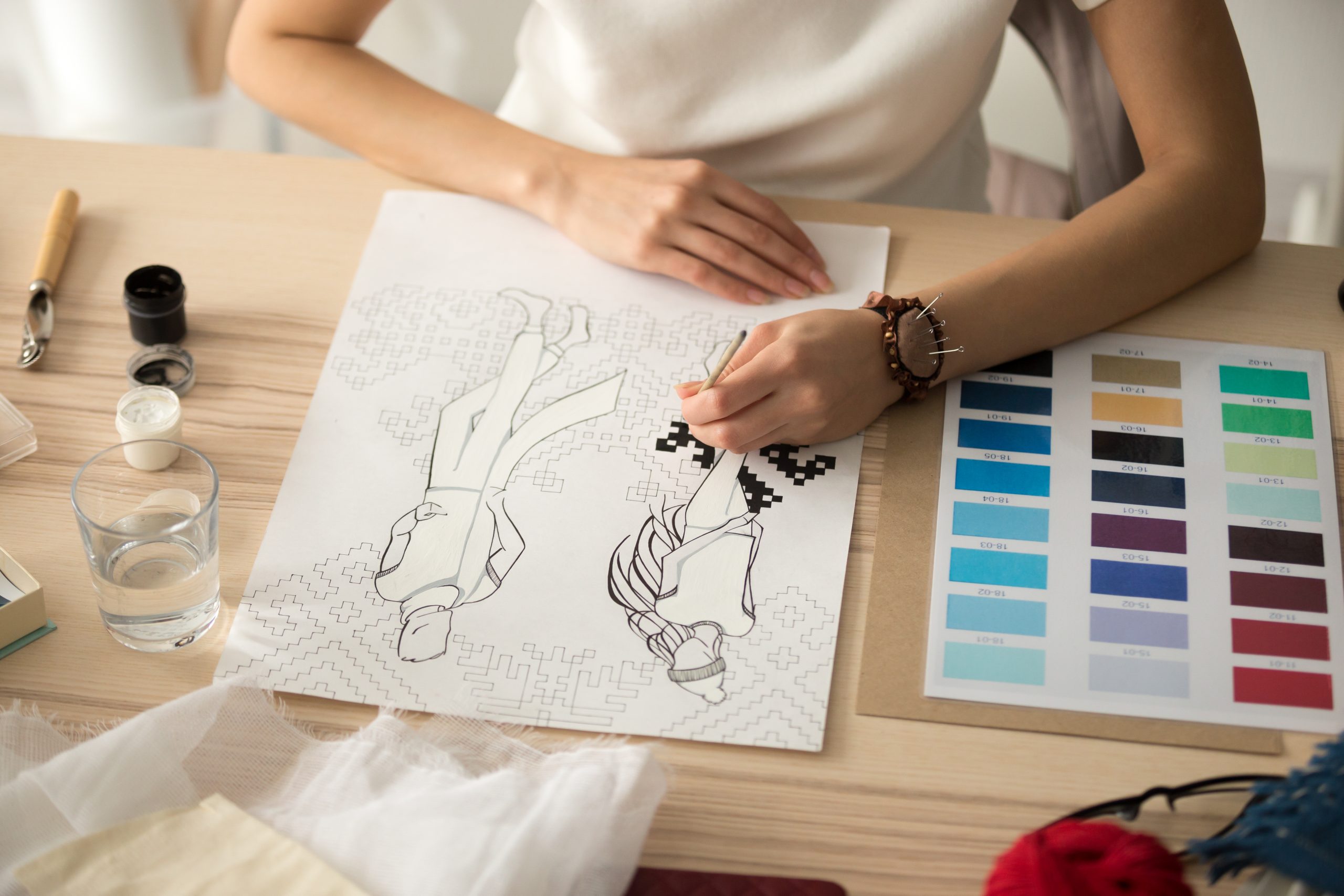Designer sketching clothing patterns with fabric swatches and color palette on a desk.